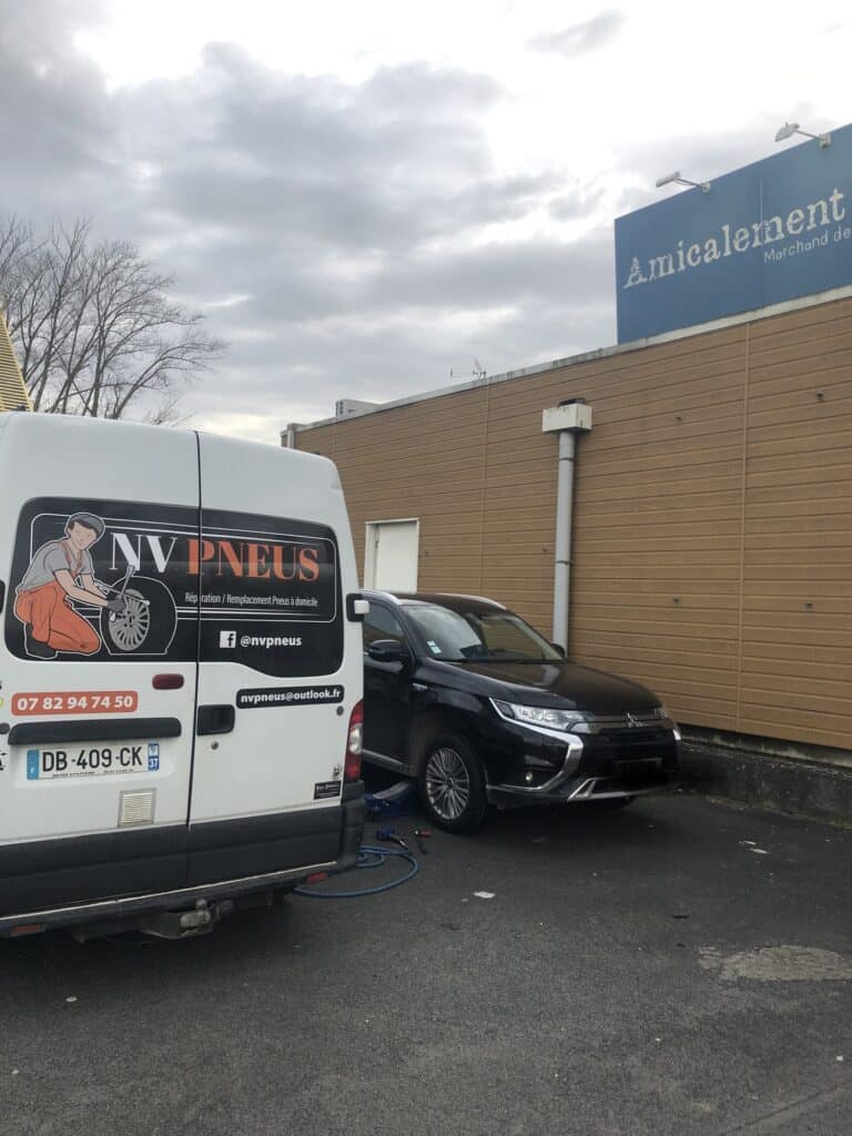 Van NV PNEUS effectuant une réparation de pneu à domicile sur un SUV noir. Texte: "Réparation / Remplacement Pneus à domicile", contacts. Panneau "Amicalement".