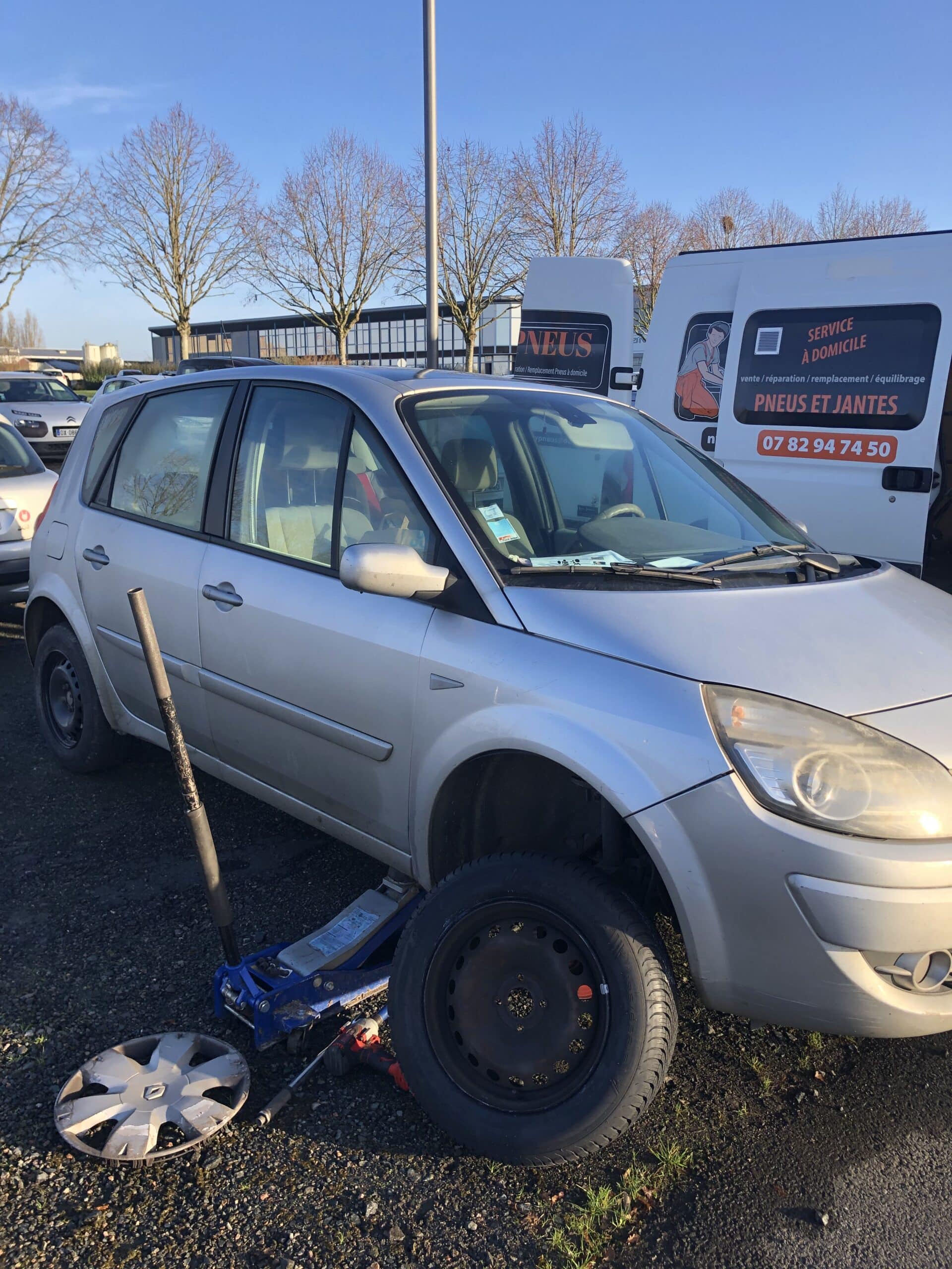 Voiture argentée sur cric avec roue démontée. Camionnette blanche "PNEUS ET JANTES SERVICE À DOMICILE 07 82 94 74 50" derrière.